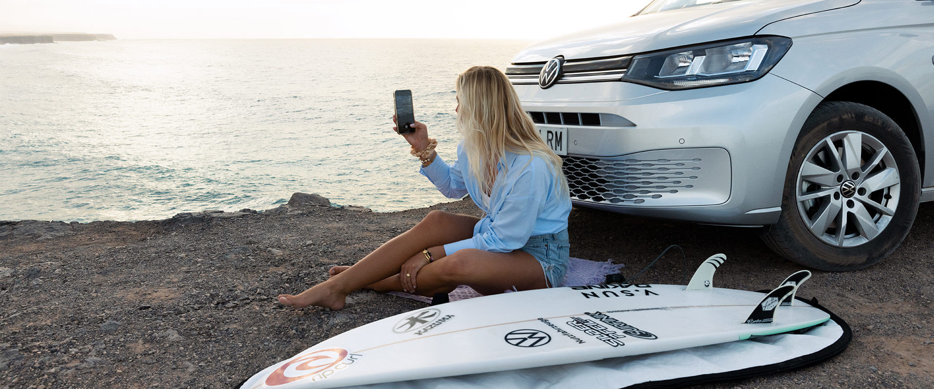 A woman sitting in front of a silver volkswagen taking a photo of the sunset